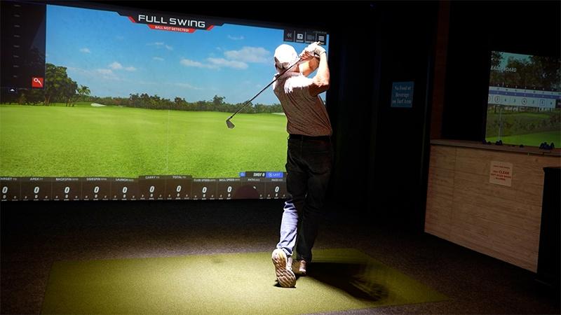 Golfer follows through on a swing from a green hitting mat in front of an indoor golf simulator screen.