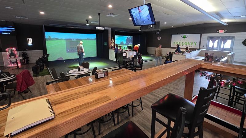 Indoor golf simulator bays with players, seating, and a wooden bar in the foreground.