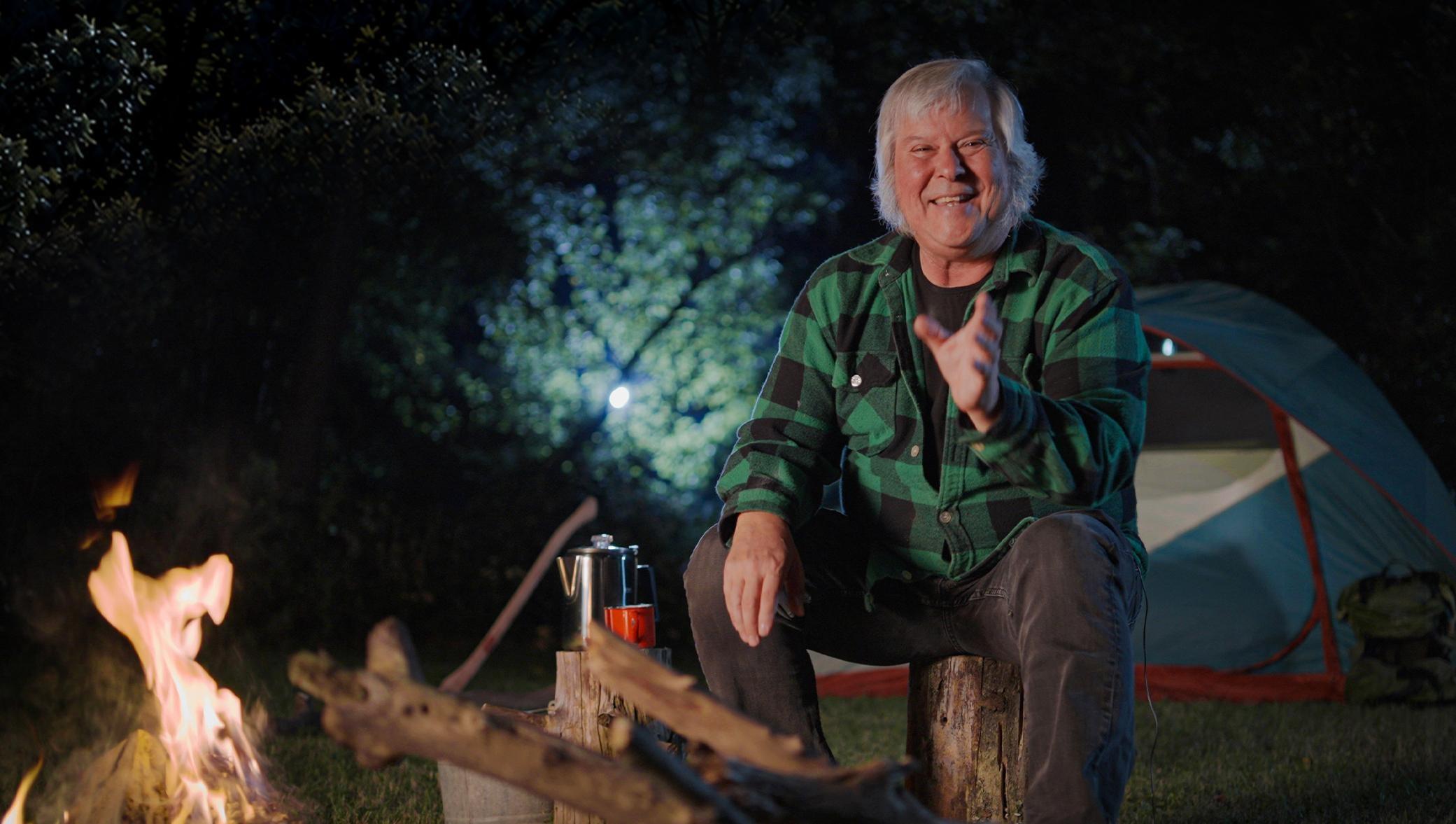 A man sits in front of a campfire at the edge of the woods holding his hand out as he tells a story