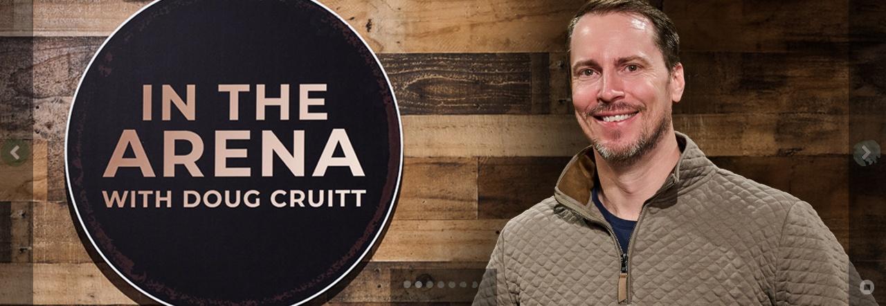 Host Doug Cruitt stands smiling next to the logo reading In The Arena with Doug Cruitt written in copper encircled over a wooden background