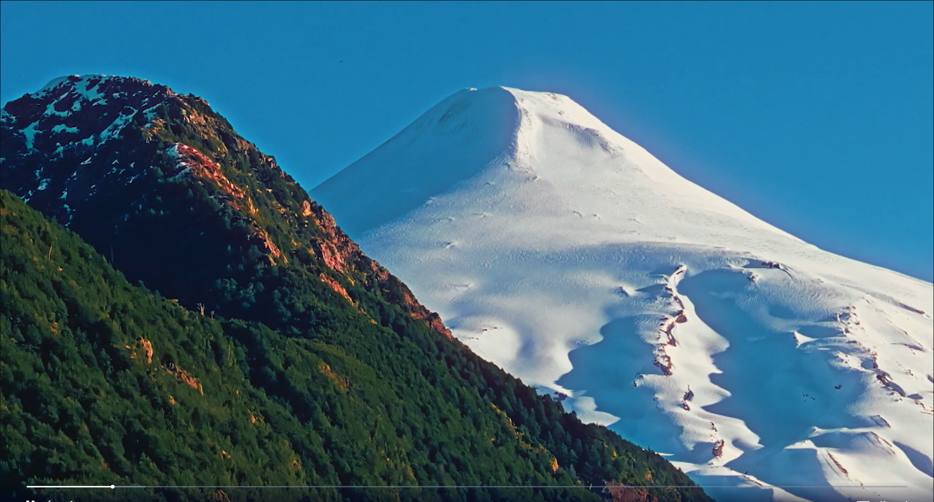 Two peaks one covered in green in the foreground, the other the snowcapped Volcano Villarica against a clear blue sky