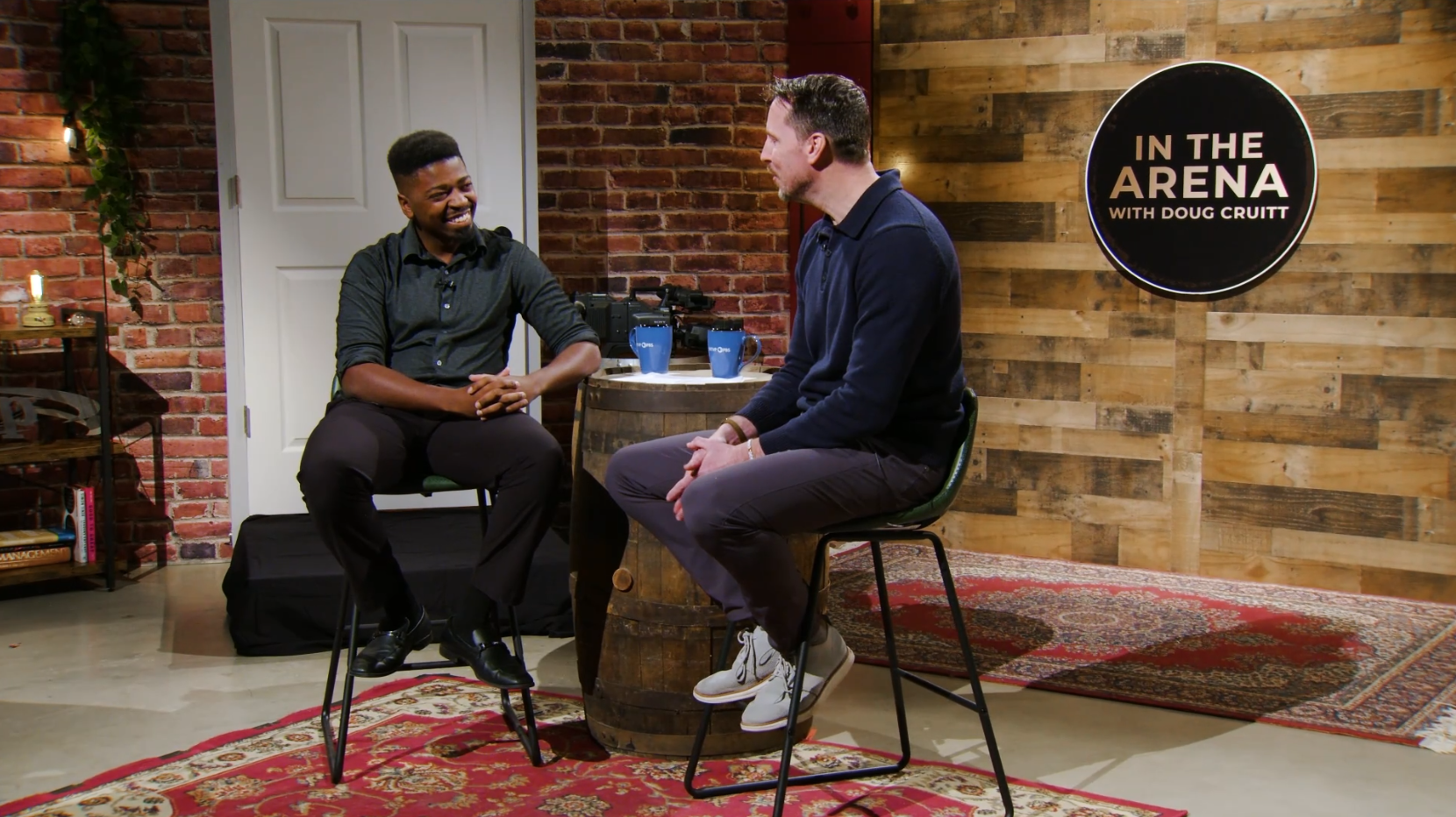 Host Doug Cruitt sits with and interviews Pierre Paul a smiling, sharply dressed, Peoria entrepreneur asking him to "tell the story behind the startup."