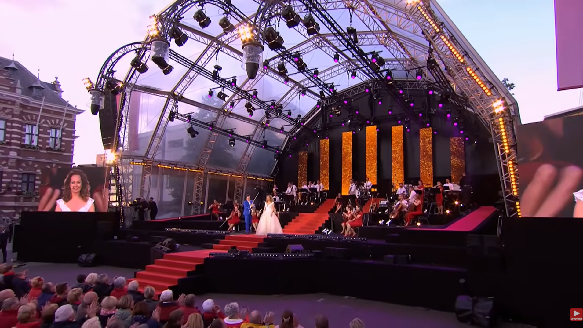 A crowd sits before a bandshell where Maestro & The European POP Orchestra play at sunset.