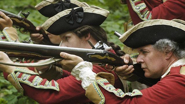 British soldiers in red coats stand in a cluster and take aim with their muskets.