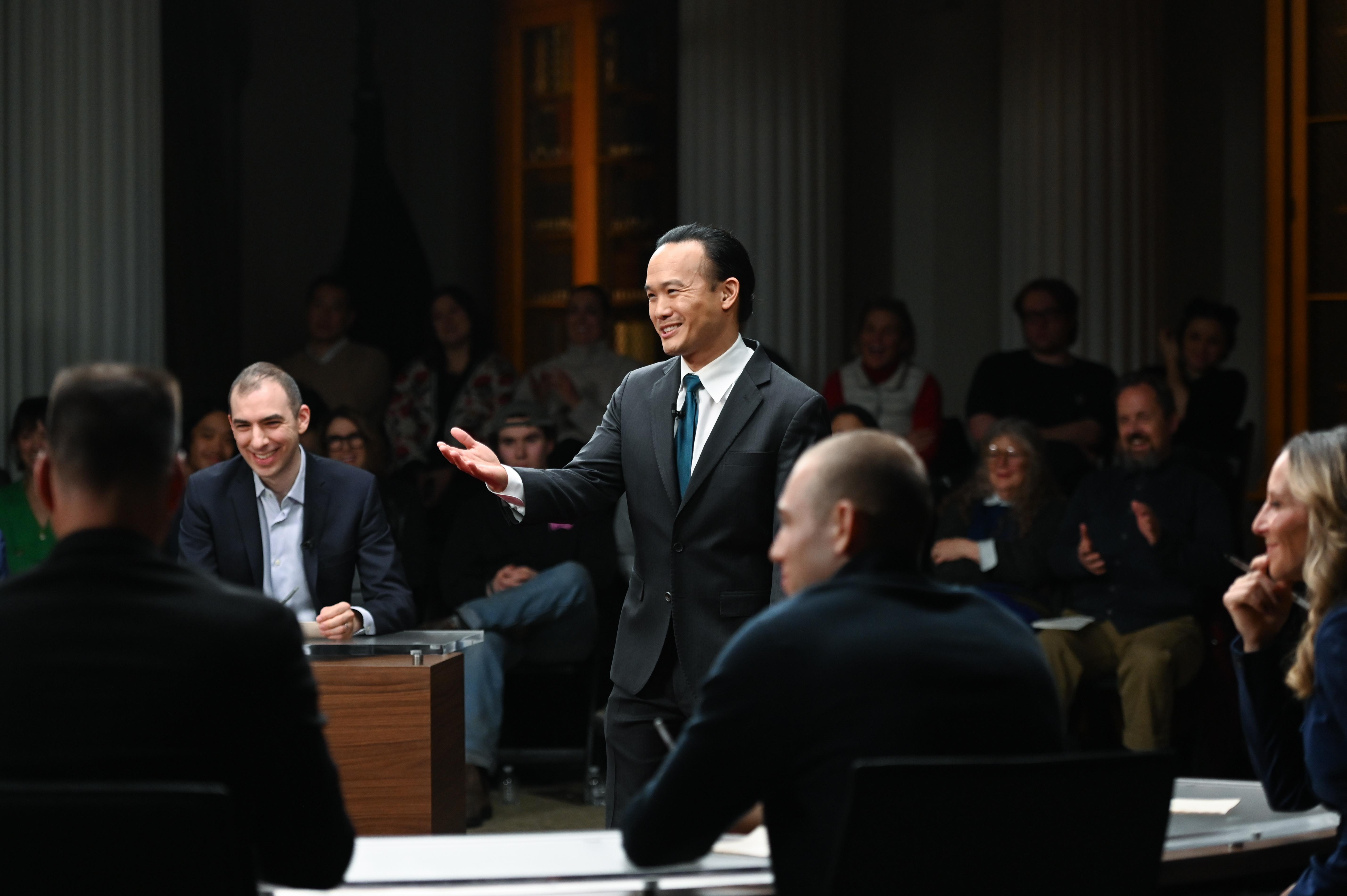 Aaron Tang stands smiling in the midst of moderating from the center of a circle of panelists who are also smiling and laughing surrounded by a live audience.