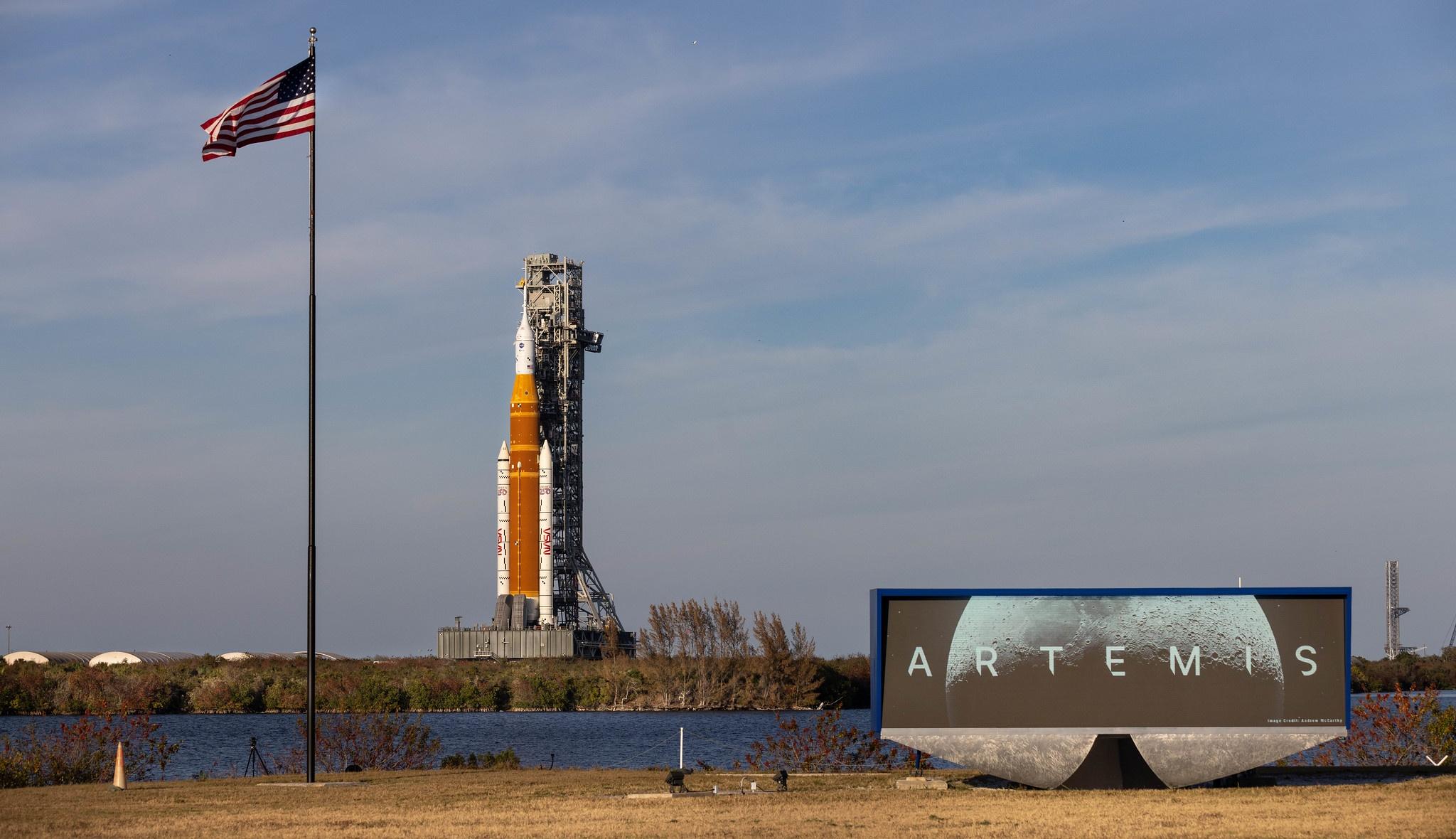 NASA's SLS rocket and Orion spacecraft tower in the distance on a sunny day with an american flag waving on the right and a countdown clock in the foreground that says "Artemis" with a lunar background.
