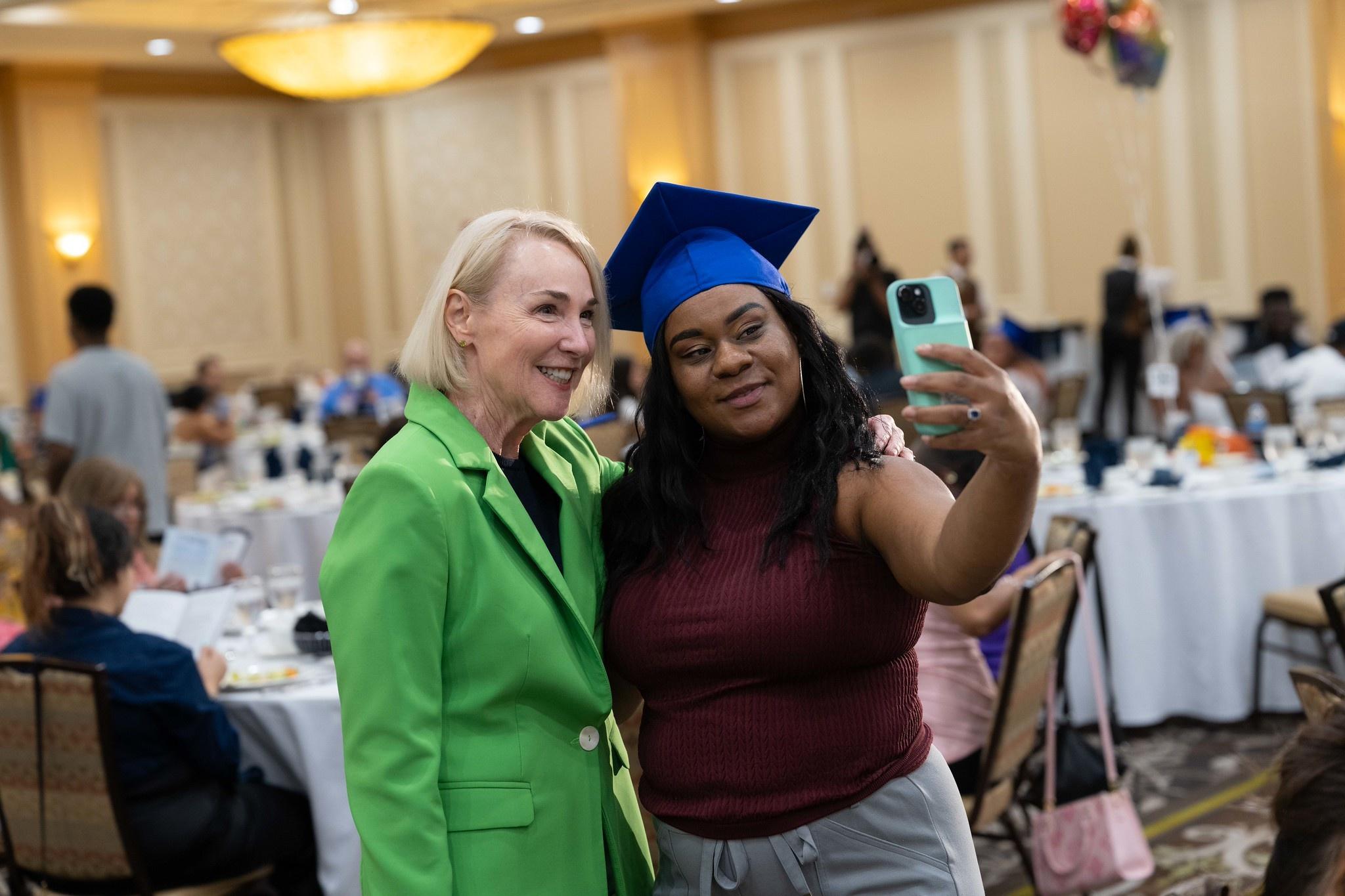 Mare Mazur wearing a green suit jacket and taking a selfie with a graduate wearing a blue cap in a hotel ballroom