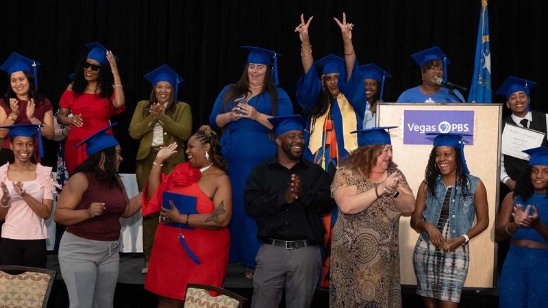 Graduates wearing blue caps and gowns celebrate onstage while standing near a Vegas PBS podium
