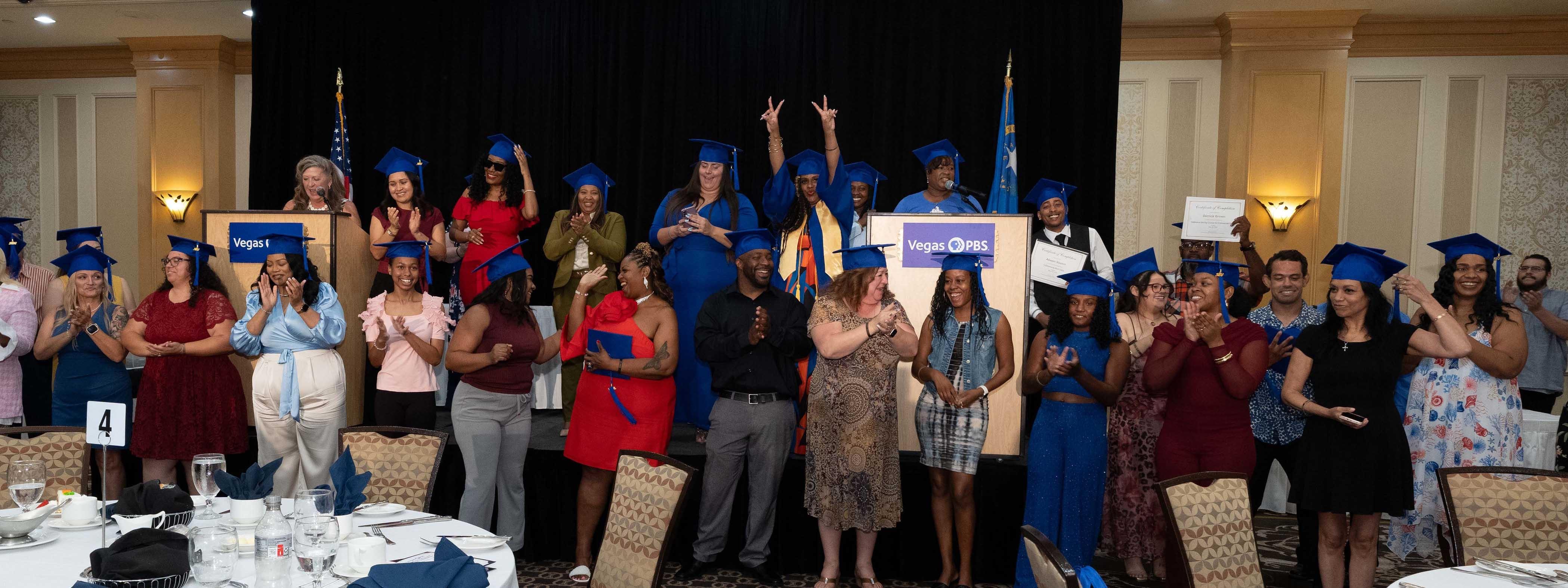 Adult graduates wearing blue caps and gowns celebrate on a stage and behind the podium in a hotel ballroom