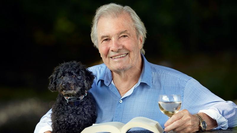 Jacques Pepin holding a glass of white white in his left hand while sitting at a table with an open book in front of him and his right arm around a curly-haired black dog
