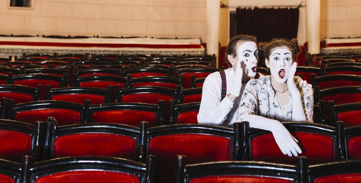 Two actors wearing white gloves and white mime makeup sit expressively in red theatre seats facing forward