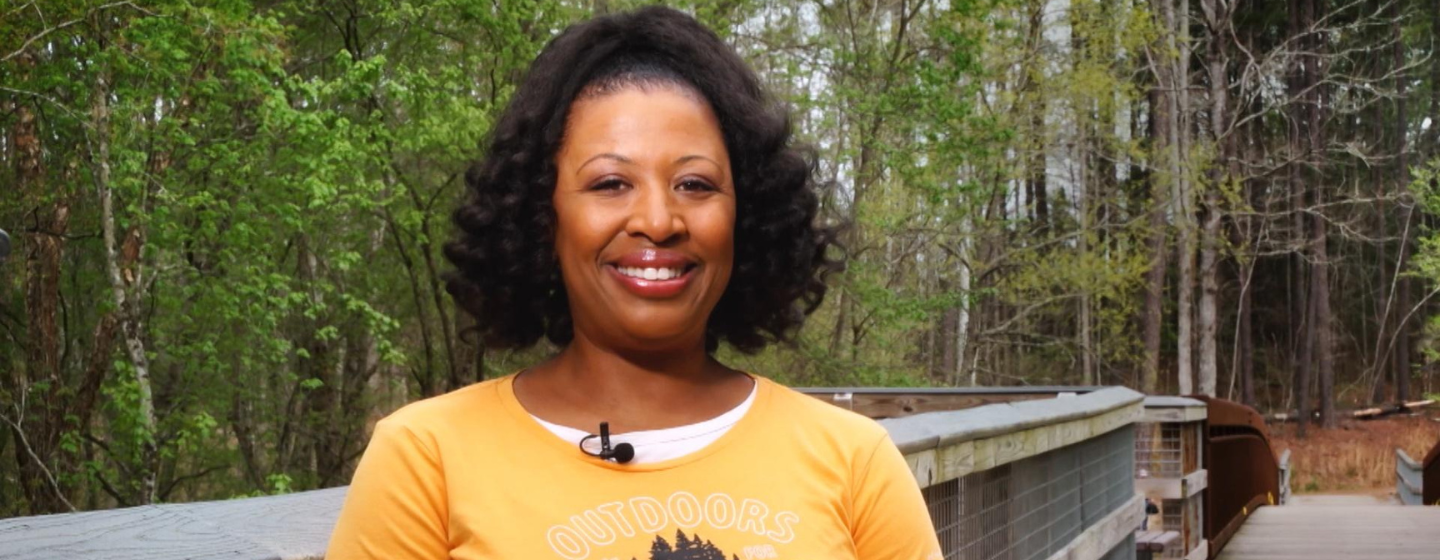 Host Deborah Holt Noel, a Black woman, stands smiling, wearing a yellow and white shirt. She stands on a wooden bridge in a green forest.