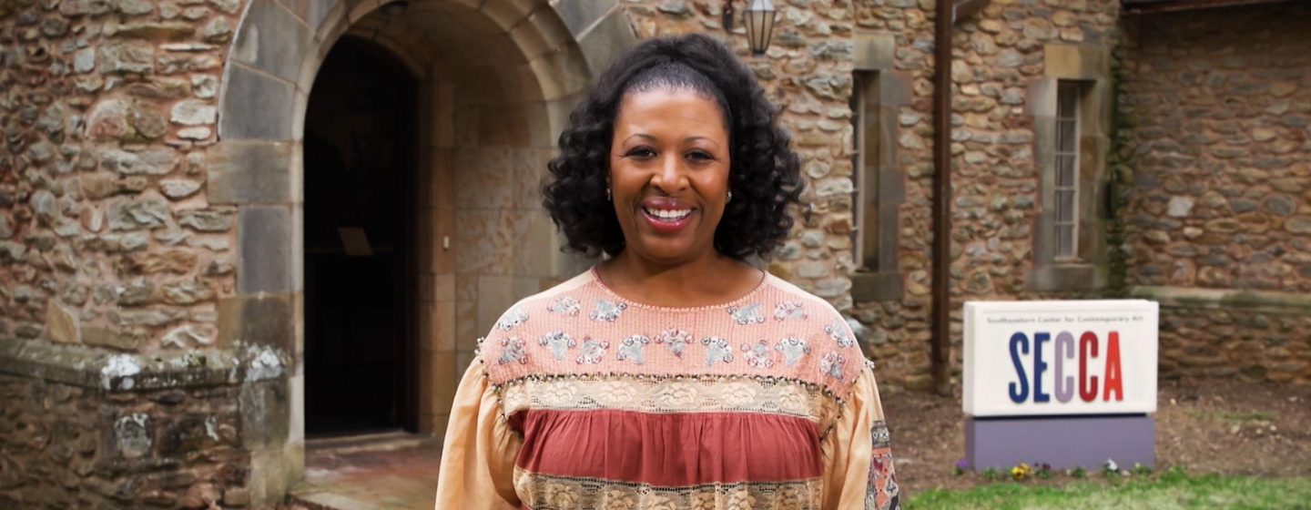 Host Deborah Holt Noel, a Black woman, stands smiling, wearing a pink and red shirt. Behind her is an old stone building and a white sign with SECCA.