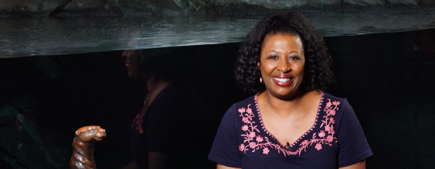 Host Deborah Holt Noel, a Black woman, is smiling, wearing a dark blue shirt with pink flowers. She is sat in front of a large, dark aquarium tank filled with water. To the left is a bronze statue of an otter standing up. 
