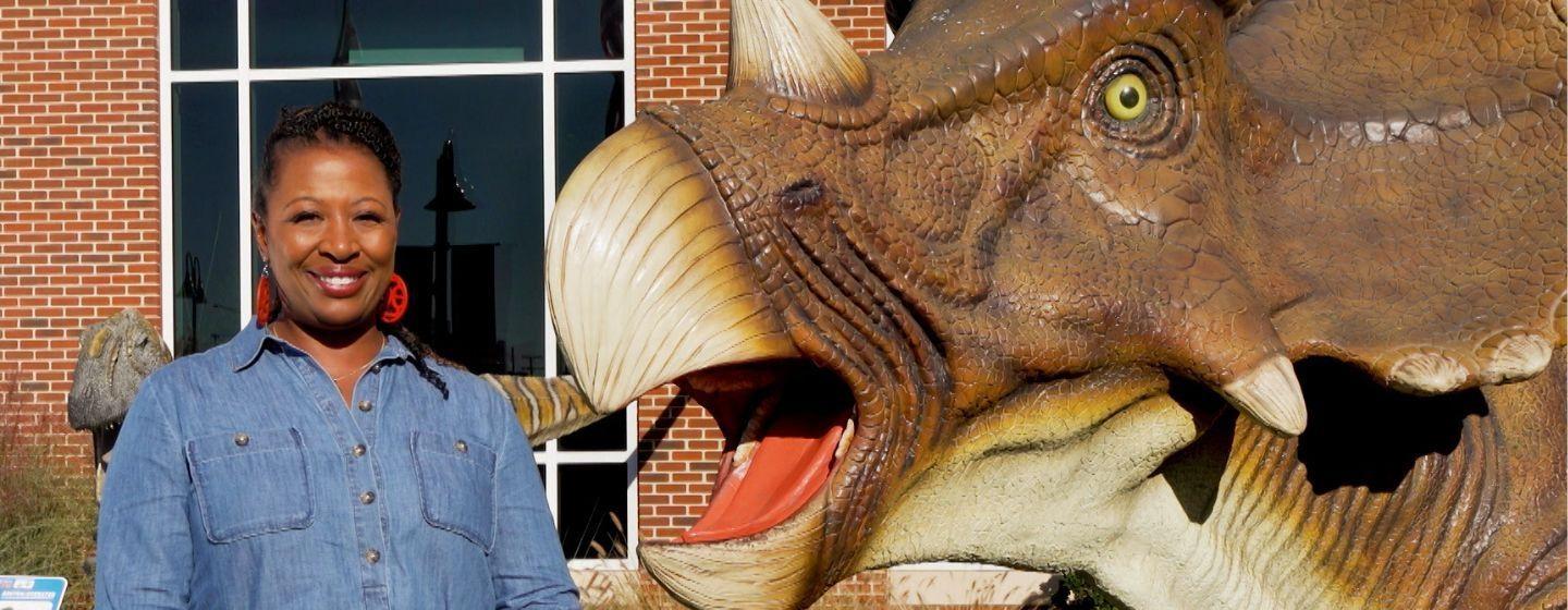 Host of NC Weekend, Deborah Holt Noel, stands next to a statue of a triceratops with it's mouth open while wearing a blue jean shirt and bright red circular earrings.