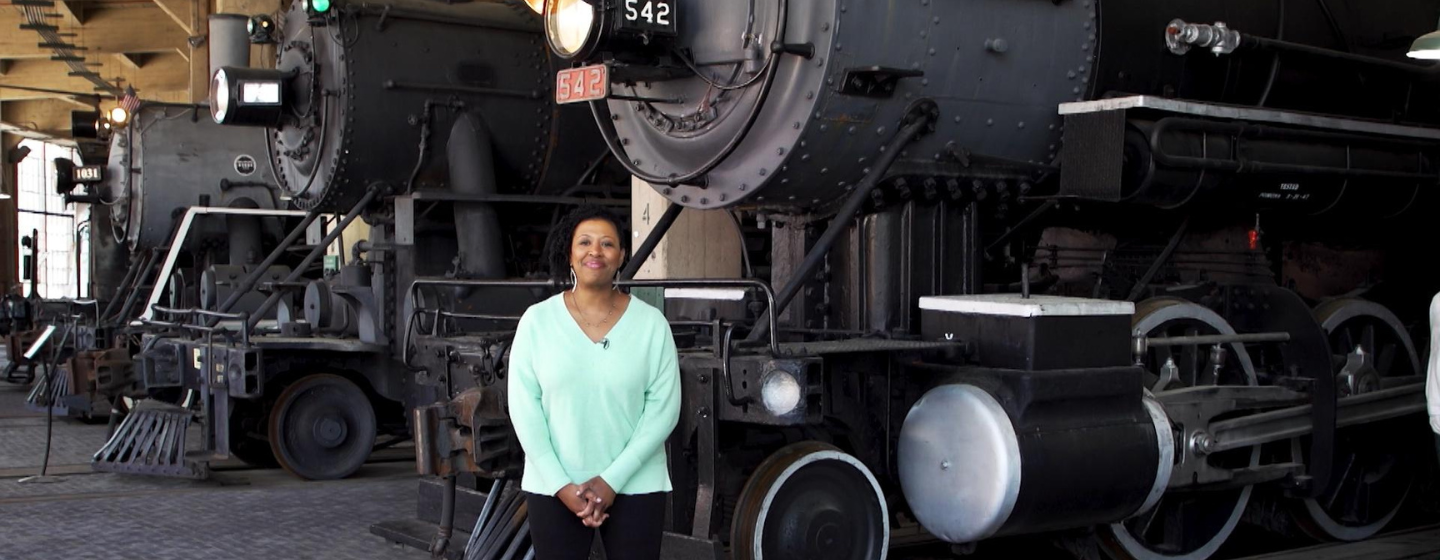 Host Deborah Holt Noel, a Black woman, stands smiling, wearing a light blue shirt and black pants. She stands in front of three large black trains.