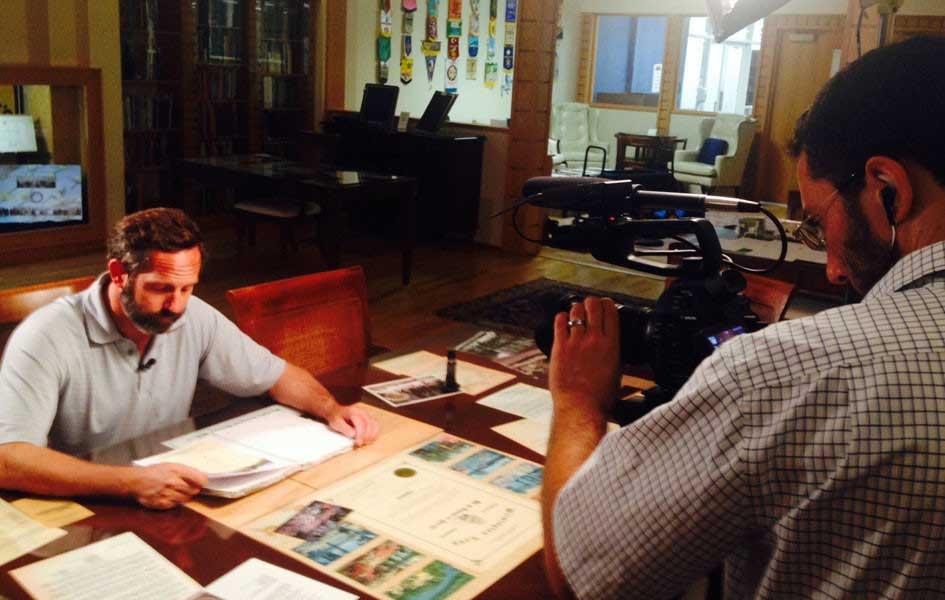 person with camera filming person sitting at table looking over old papers and files
