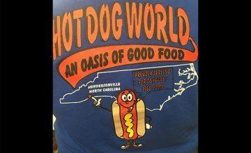 Photo of the back of a "Hot Dog World" blue t-shirt. Featuring the slogan "An oasis of good food" on a red banner.