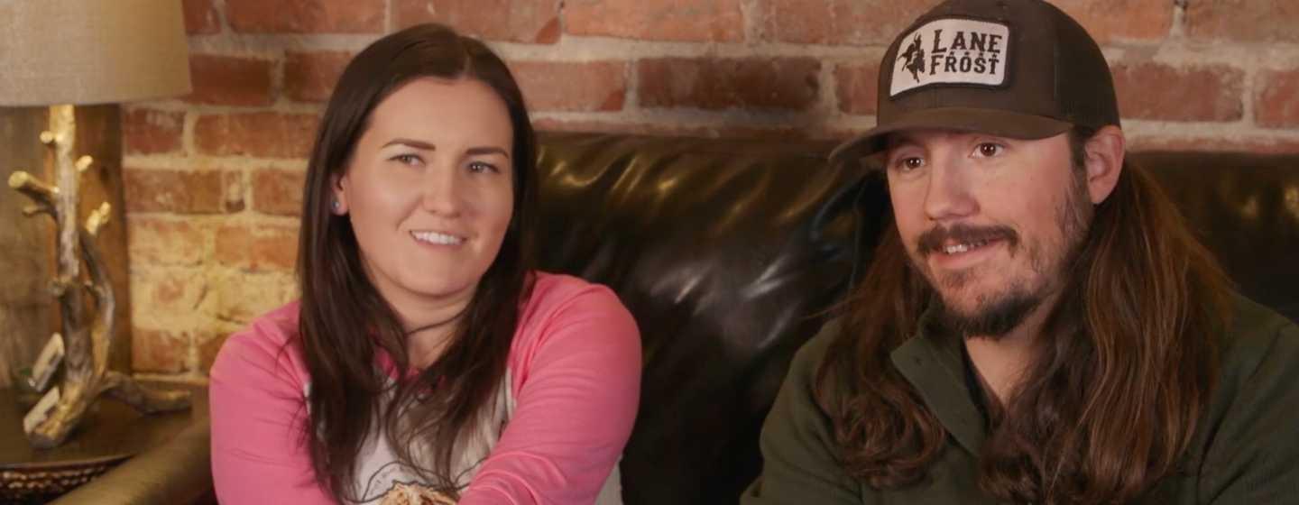 Emily and Adam Hopson sit on a dark couch in front of a brick wall.