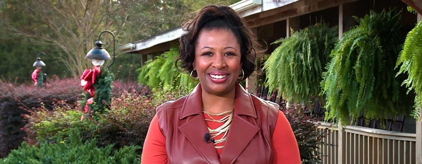 Deborah Holt Noel, wearing a red sweater and dark red vest, smiles at the camera with winter greenery in the background