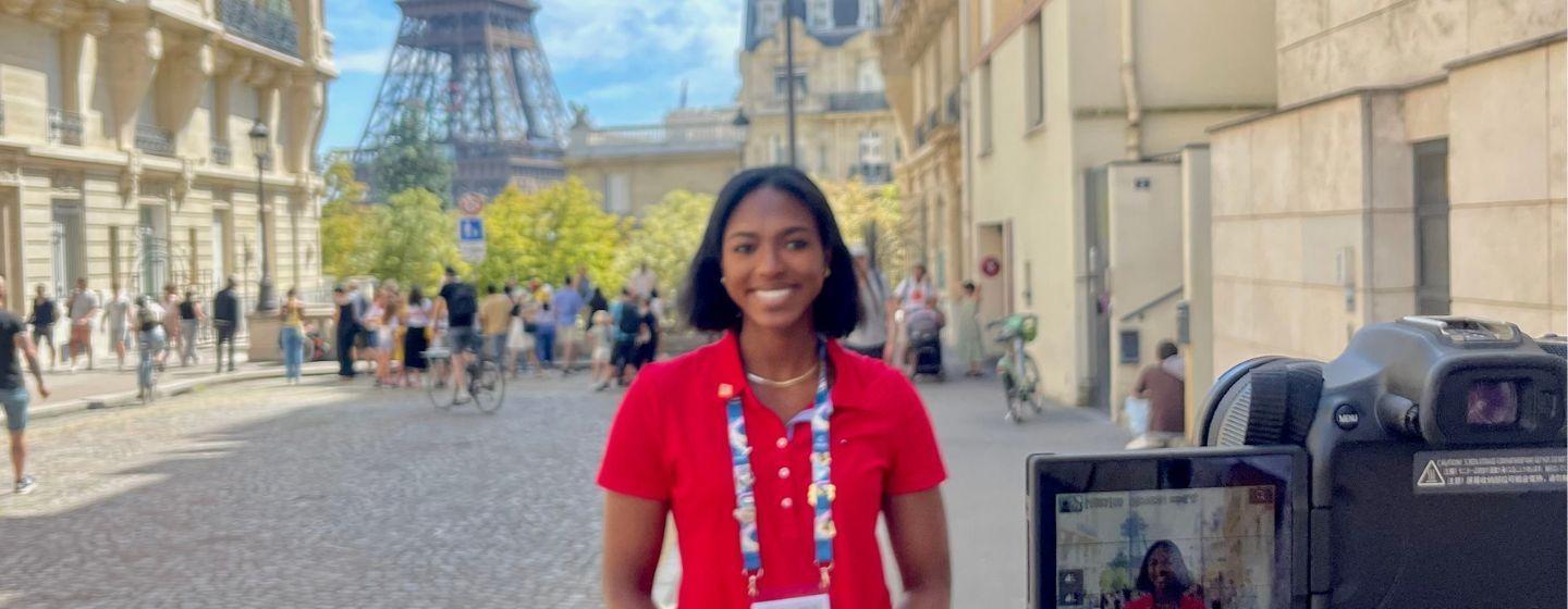 Jasmine Baker poses in front of a camera and with the Eiffel Tower in the background