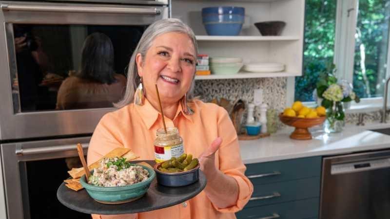 Sheri Castle smiles brightly and holds up a platter of crackers, ham salad spread, pickles and mustard in a kitchen