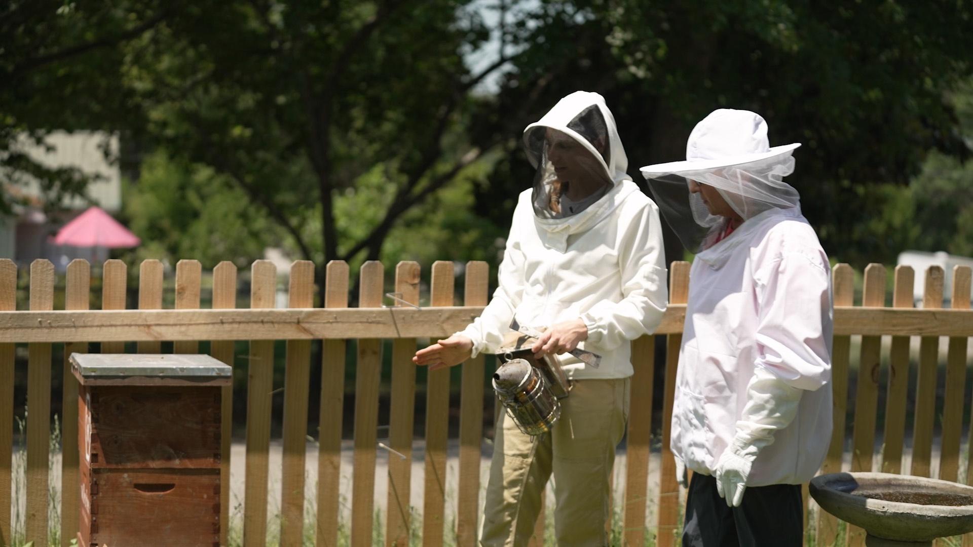 Mary Garrison and Sheri Castle in beekeeping suits standing near a handmade bee house.