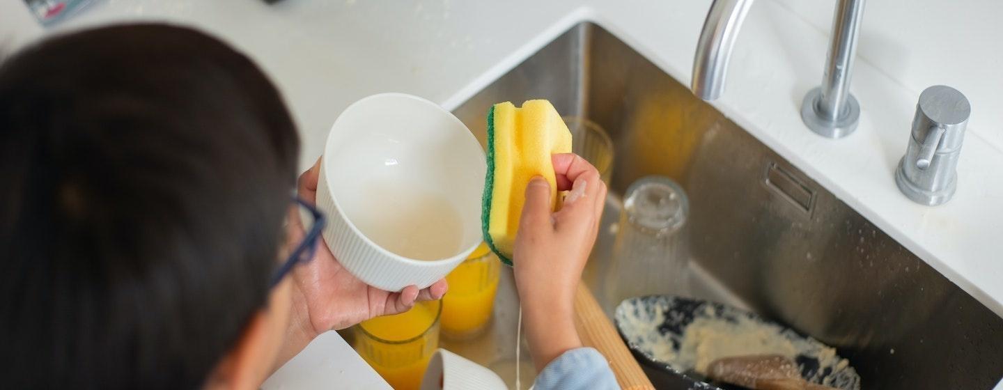 overhead man cleaning small white bowl with yellow and green wet sponge over kitchen sink with dirty dishes inside below 