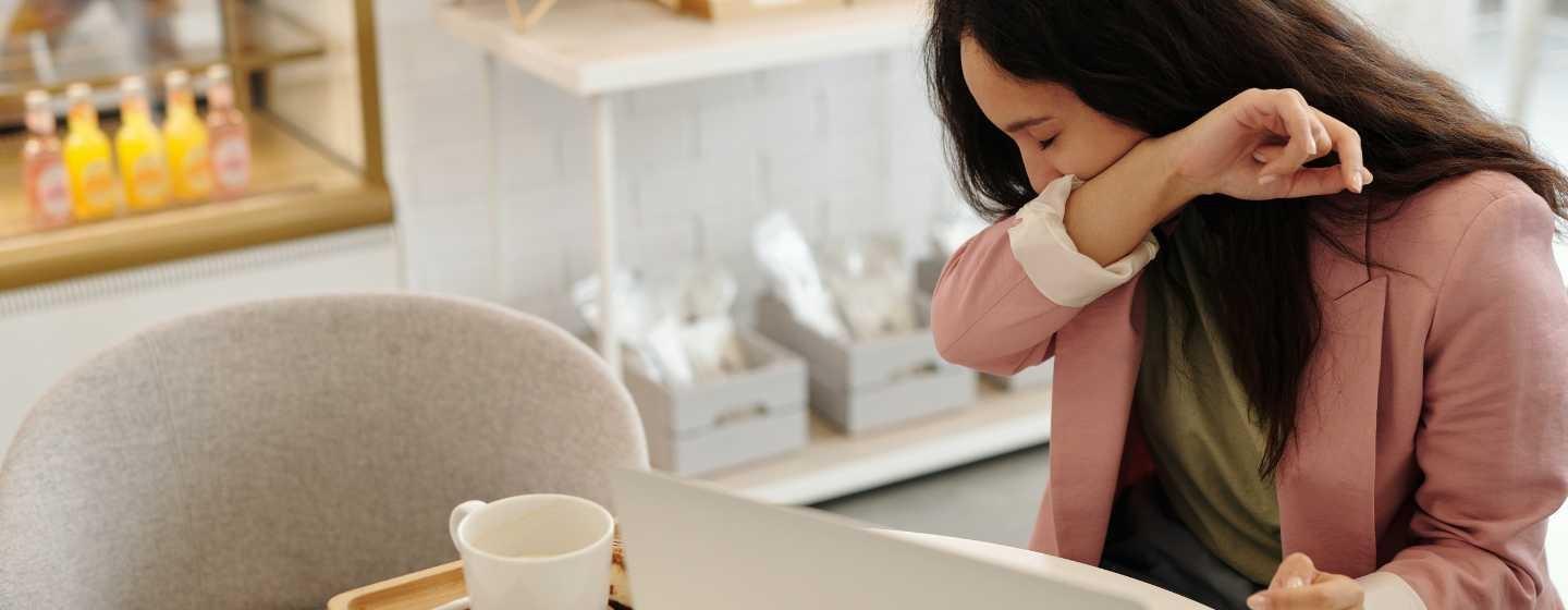 A woman wearing an olive green shirt and a muted pink blazer sits at a table with a coffee cup and laptop open, leaning her face into her right elbow to sneeze