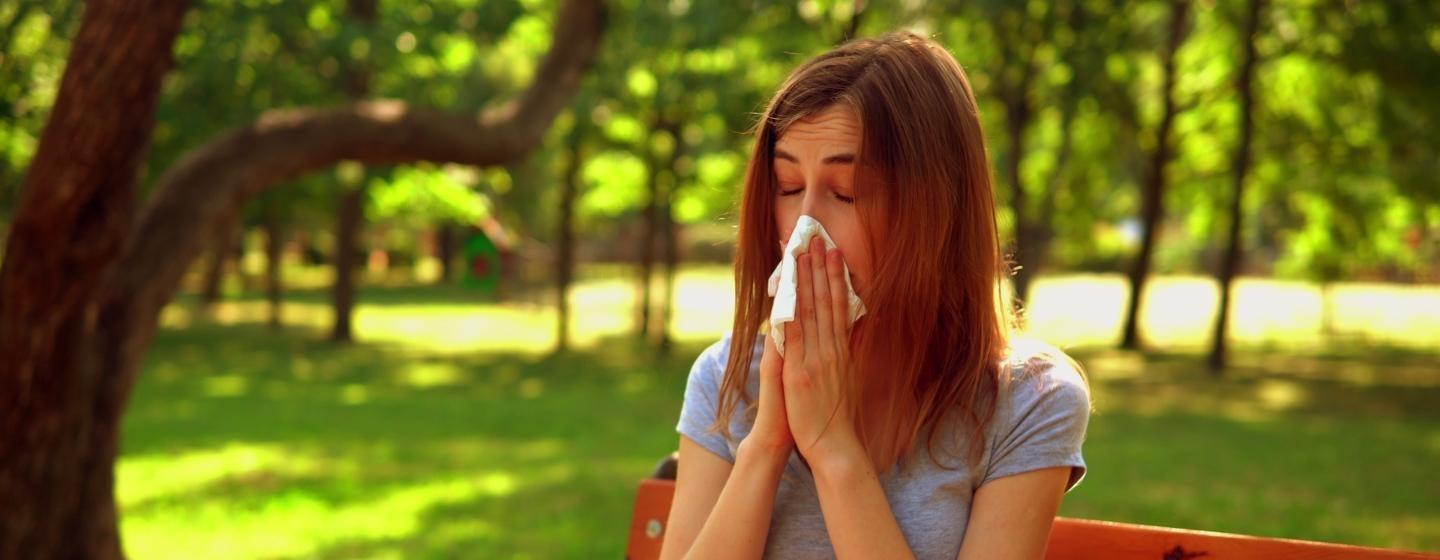 A woman blowing her nose while sitting on a bench in the park.