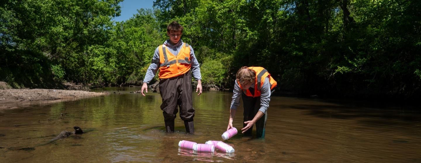 Two people wearing orange safety vests in a creek examining pink plastic bottles.