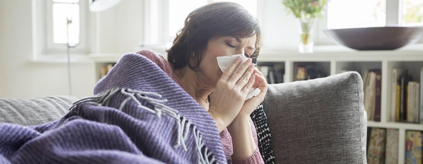 woman lying under blanket on couch sneezing into tissue in living room morning light coming through windows