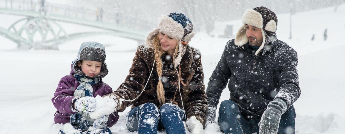 A child and two adults dressed in lots of winter clothing sit in a field of snow while flurries fall.