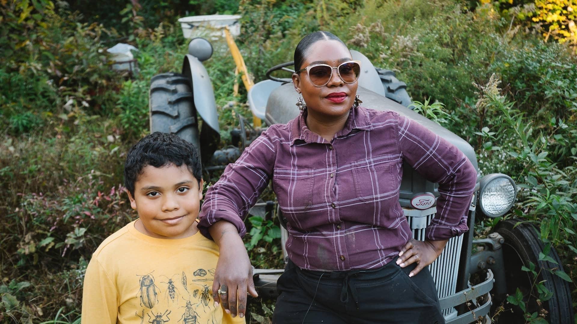 farmer woman and kid standing in front of old tractor with hand on kid's shoulder standing in dense undergrowth