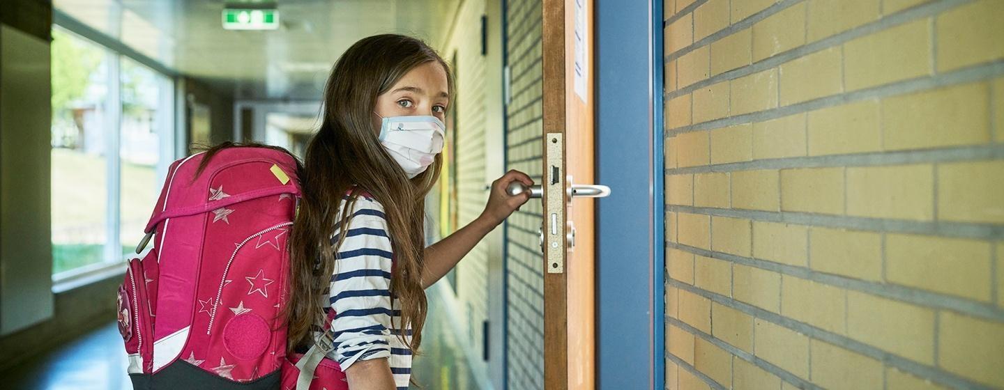 masked girl with pink backback from side at school classroom opening door