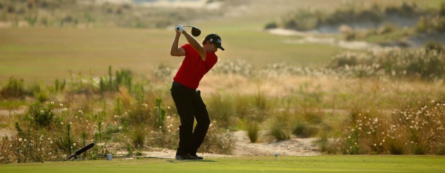 A golfer in a red polo shirt, black pants and a black baseball cap swings with a driver.