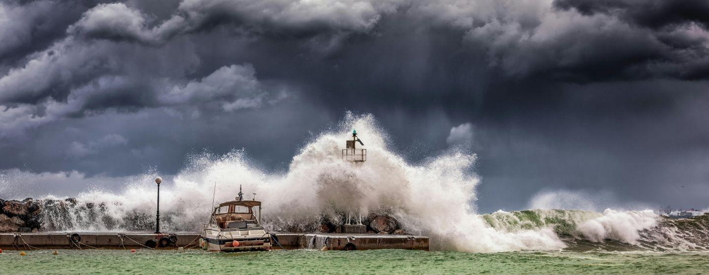 A large wave from the green sea crashing over a dock with boats on it. The sky is full of dark gray storm clouds.
