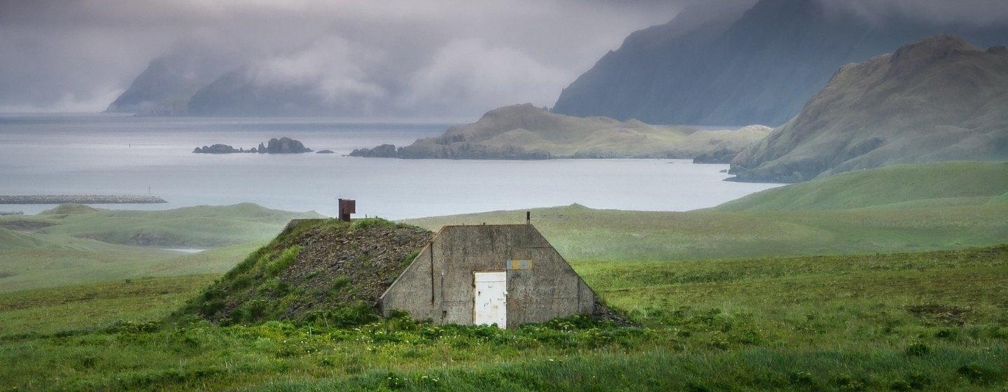A grassy field with the entrance to a WWII bunker in the middle and a mountainous coast in the background.