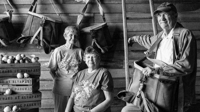 A black-and-white photograph with three older white people in them. Two women and one man, all in comfortable clothes for outdoor work (T-shirts and jeans); the man wears a hat. They are posing in front of a horizontally slated wooden wall of what appears to be an older building. There are apple bags hanging on the wall, and the man holds one of them on his shoulders. Wooden crates of apples repeatedly stamped with the word “Altapass” are stacked to one side of the frame.