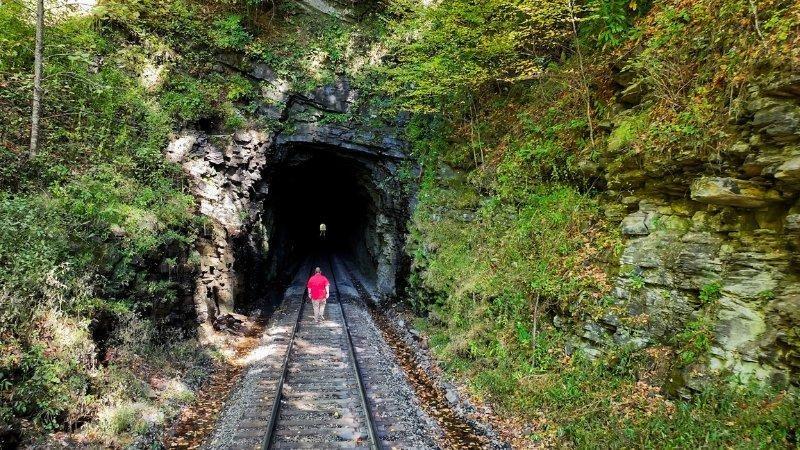 A man with blond hair in a bright red shirt walks away from the camera down railroad tracks that disappear into a dark tunnel in a rocky mountainside. The other end of the tunnel is just visible, and the man is small in perspective in the photograph. The gray, rocky walls are covered in green vegetation and dappled with sunlight.