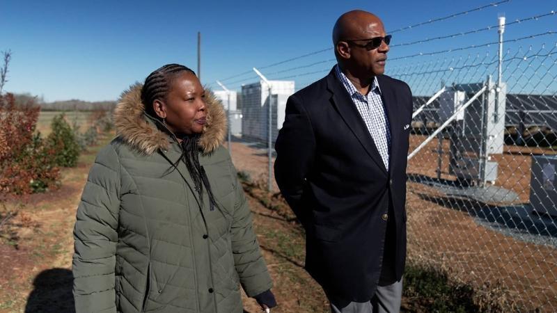 A man and woman walk along a fence line. They are looking at what is inside the fence, a collection of solar panels and metal boxes housing batteries and electrical components.
