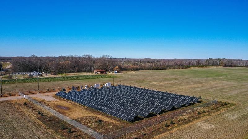 An array of seven long solar panels in Pendleton, North Carolina, is surrounded by slightly green agricultural fields. In the background, a small house stands with rows of green crops nearby.