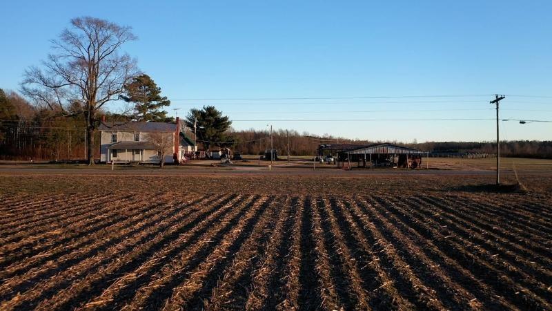 View looking down tilled rows in an agricultural field at an old white farmhouse and some farm outbuildings. A newer-looking house is in the background. Farther into the background in the same field, you can see a small installation of solar panels.
