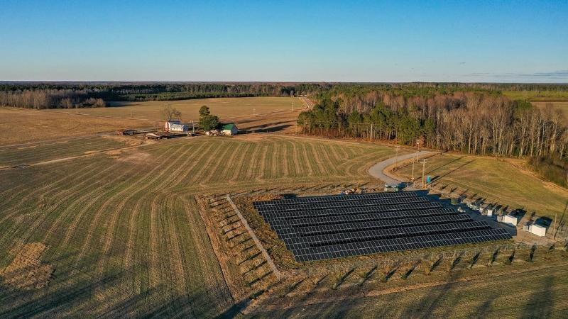 An array of seven long solar panels surrounded by tilled agricultural fields. A cluster of buildings, including several houses, sits nearby.