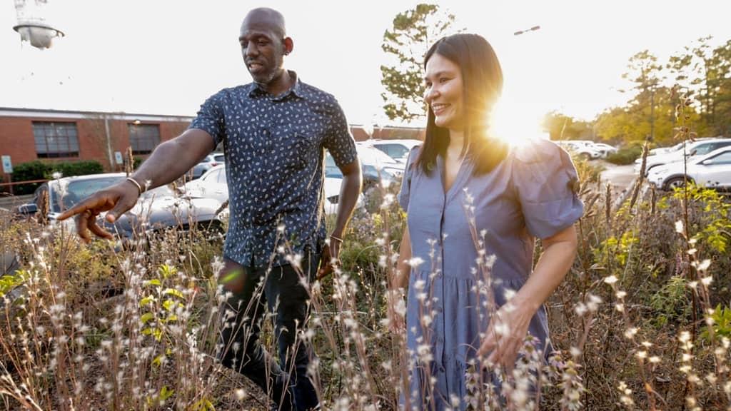 A man and woman wearing a dark blue shirt and lighter silvery blue dress stand amidst grasses and plants that reach almost to their shoulders in the evening light. The setting sun peaks over the woman’s shoulder.