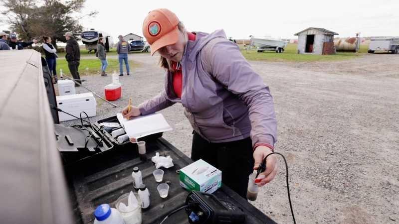 A woman in a purple raincoat and faded orange cap stands on a gravel road at the back of a truck, working with scientific equipment laid out on the tailgate.