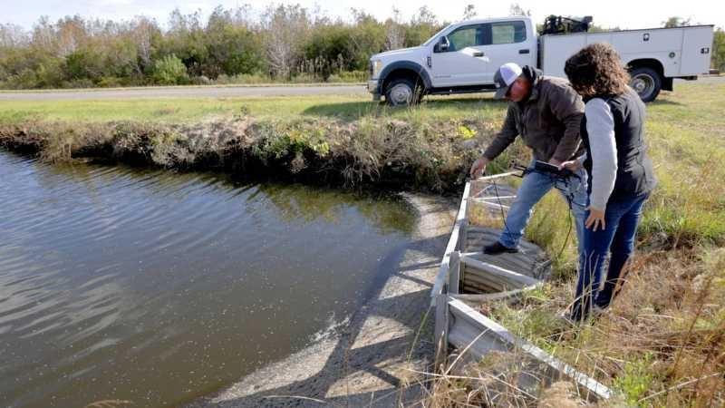 Two farmers stand above a corrugated metal structure on a ditch filled with water, holding a piece of equipment with an attached cable that they’ve dropped into the water of the ditch.