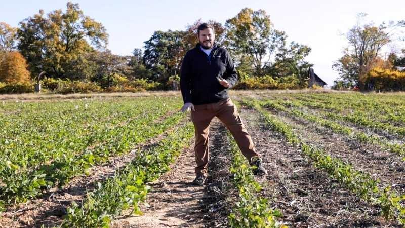 A man in a black fleece jacket and tan pants stands straddling a row of green soybean plants in a field of soybean plants. His right foot is lower than his left because the ground is lower on one side of the row of plants than the other.
