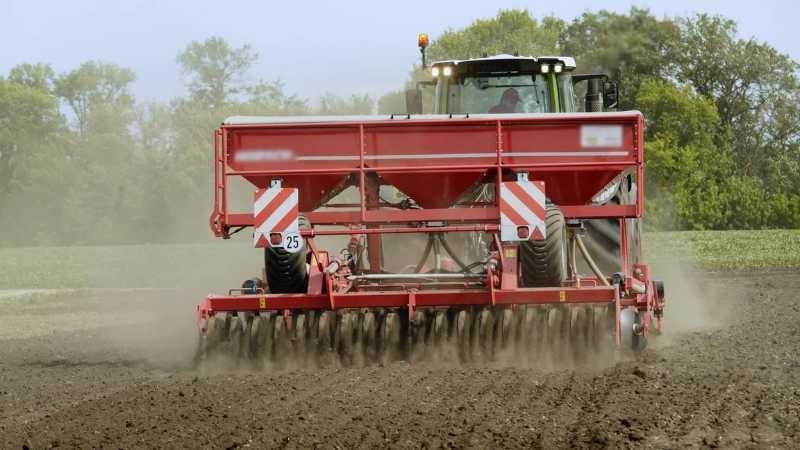 A piece of farm equipment is pulling a red tilling attachment away from the viewer, and the tiller has round discs that are touching the soil. The soil is dark brown, and there’s a slight cloud of dust around the machine.