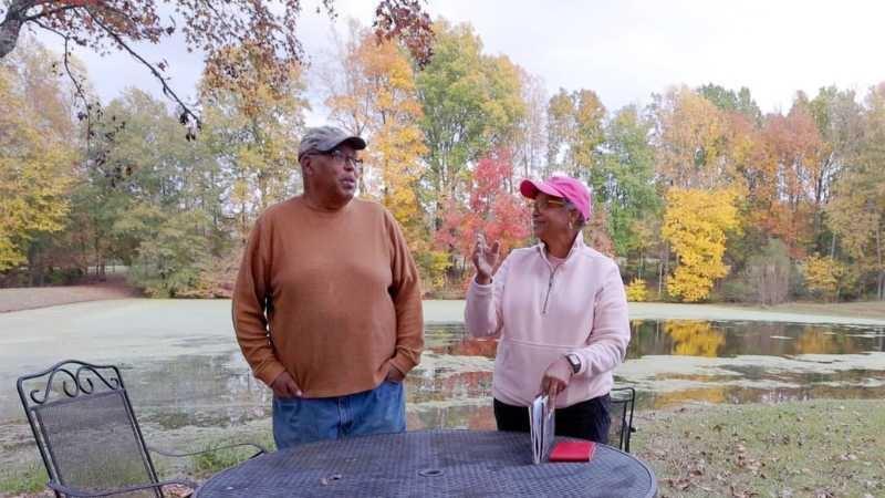Two people are standing behind a black metal table outdoors in front of a large pond surrounded by trees with fall leaf colors of bright red and yellow.